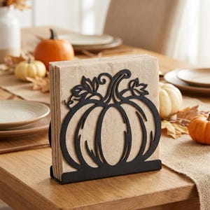 May include: A black metal napkin holder featuring a cutout pumpkin design on the front panel. The holder is filled with a stack of light tan paper napkins. The pumpkin silhouette includes detailed stems and leaves at the top of the metal frame.