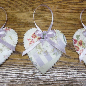 May include: Three lavender and white fabric heart ornaments with a pearl trim and a lavender satin ribbon bow with a small flower accent.