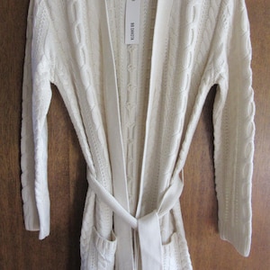 May include: A cream-colored cable knit cardigan with a self-tie belt and pockets. The cardigan has a long, relaxed fit and is made of a soft, comfortable material. The tag on the cardigan reads "BB Dakota".