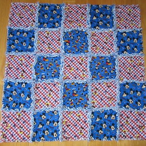 May include: A blue and red patchwork quilt with a Mickey Mouse pattern. The quilt is made of squares of different fabrics, some with a gingham pattern and some with a Mickey Mouse print. The quilt is edged with a white ruffle.