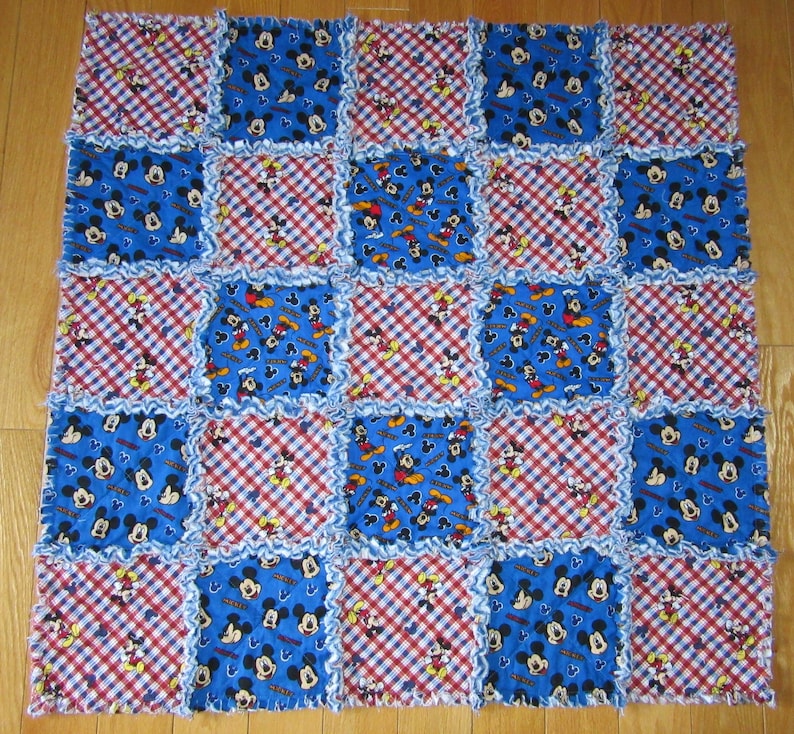 May include: A blue and red patchwork quilt with a Mickey Mouse pattern. The quilt is made of squares of different fabrics, some with a gingham pattern and some with a Mickey Mouse print. The quilt is edged with a white ruffle.
