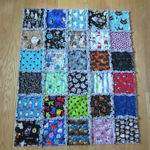 May include: A rectangular rag quilt composed of thirty square fabric patches featuring various cat-themed patterns in colours including black, blue, green, red, and purple. Each square is joined with exposed, frayed seams, creating a textured grid.