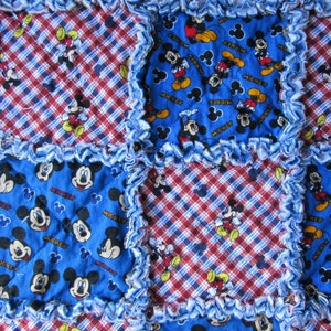 May include: A blue and red patchwork quilt with a Mickey Mouse print. The quilt is made of squares of different fabrics, some with a plaid pattern and some with a Mickey Mouse print. The quilt is edged with a blue ruffle.