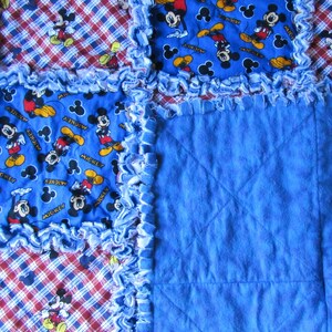 May include: A blue and red plaid fabric quilt with a blue fabric square in the center. The quilt is decorated with a blue fabric with a Mickey Mouse print. The fabric is frayed around the edges.