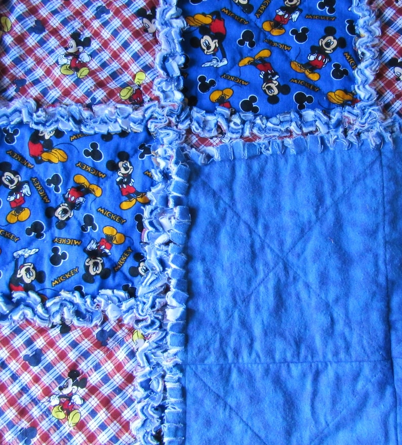 May include: A blue and red plaid fabric quilt with a blue fabric square in the center. The quilt is decorated with a blue fabric with a Mickey Mouse print. The fabric is frayed around the edges.