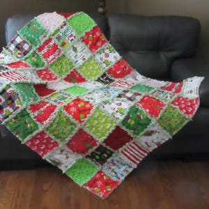 May include: A handmade Christmas patchwork quilt with a variety of festive patterns. The quilt features red, green, and white squares with holiday designs, including stripes, snowflakes, and cartoon characters. The quilt is draped over a black couch.