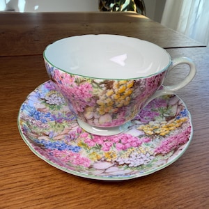 Peut inclure: Un ensemble tasse et soucoupe en porcelaine orné d'un motif floral multicolore vif avec des grappes de fleurs roses, jaunes et bleues. La tasse présente un bord festonné avec un fin liseré vert, une anse courbée et un intérieur blanc.