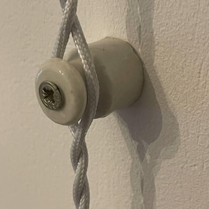 May include: A white ceramic wall insulator mounted to a surface, featuring a central metal screw. A white braided fabric cord is looped around the cylindrical body of the insulator, which is positioned vertically against the flat wall surface.
