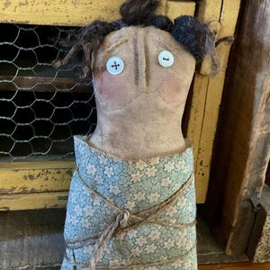 May include: A handmade doll with a tan fabric body, button eyes, and a floral patterned dress. The doll's hair is dark brown, and it has rosy cheeks. The dress is tied with twine. The doll is a folk art piece.