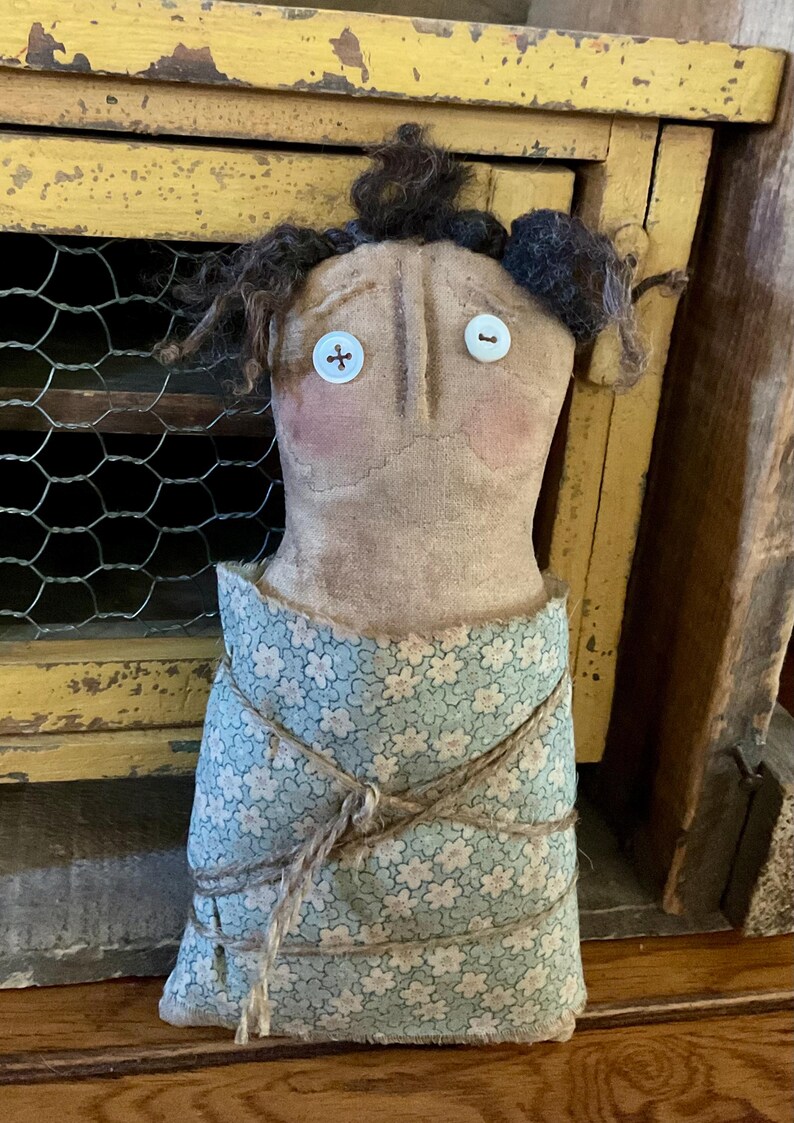 May include: A handmade doll with a tan fabric body, button eyes, and a floral patterned dress. The doll's hair is dark brown, and it has rosy cheeks. The dress is tied with twine. The doll is a folk art piece.