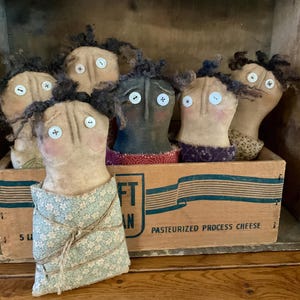 May include: A collection of handmade folk art dolls with button eyes and yarn hair, displayed in a vintage wooden box. The dolls have painted faces and are dressed in various fabrics. The box has text that reads "PASTEURIZED PROCESS CHEESE".