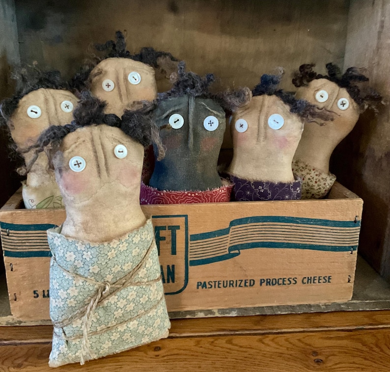 May include: A collection of handmade folk art dolls with button eyes and yarn hair, displayed in a vintage wooden box. The dolls have painted faces and are dressed in various fabrics. The box has text that reads "PASTEURIZED PROCESS CHEESE".