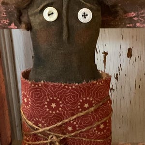 May include: A folk art doll with a dark brown head, button eyes, and curly hair. The doll's body is wrapped in a red fabric with a white star pattern, tied with twine. The doll has a rustic, handmade appearance.
