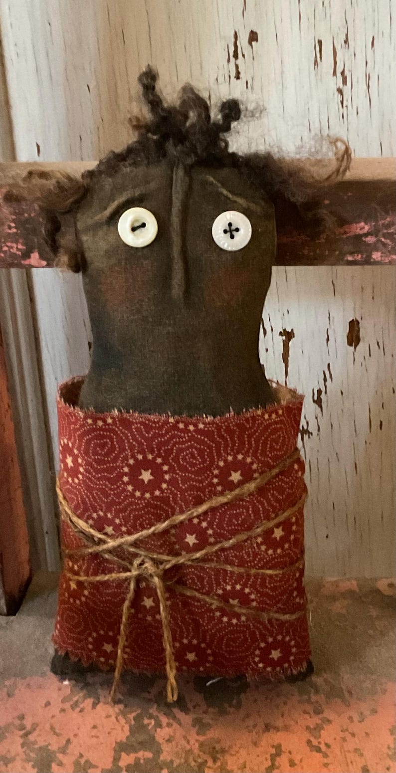 May include: A folk art doll with a dark brown head, button eyes, and curly hair. The doll's body is wrapped in a red fabric with a white star pattern, tied with twine. The doll has a rustic, handmade appearance.