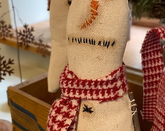 May include: Handmade snowman doll with a burlap body, embroidered face, and a red and white patterned scarf. The snowman has twig arms and black button accents. A festive winter decoration.