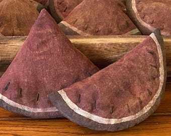 May include: Close-up of several watermelon-shaped fabric ornaments. The ornaments are primarily burgundy with a white stripe and dark brown rind. Black stitches represent seeds. The ornaments are arranged in a wooden tray.