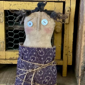 May include: A handmade doll with a tan fabric face, button eyes, and curly black hair. The doll has rosy cheeks and is wrapped in a dark purple fabric with a white floral pattern, tied with twine. The doll is a folk art style.