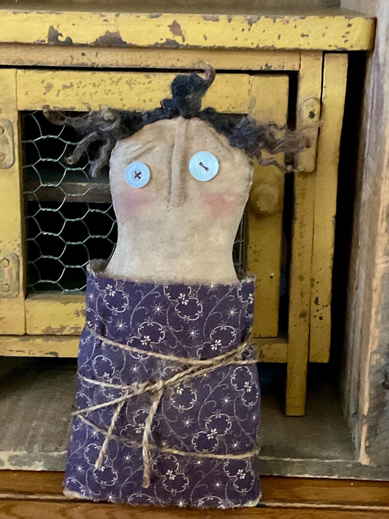 May include: A handmade doll with a tan fabric face, button eyes, and curly black hair. The doll has rosy cheeks and is wrapped in a dark purple fabric with a white floral pattern, tied with twine. The doll is a folk art style.