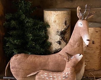 May include: A brown fabric deer figurine standing next to a smaller brown fabric fawn figurine. The deer has small twig antlers and a light-colored muzzle. The fawn features white painted spots along its back. Both figures have stitched details.
