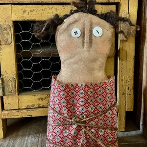 May include: A handmade fabric doll with a tan face, button eyes, and dark yarn hair. The doll wears a red and blue patterned dress tied with twine. The doll is displayed in front of a distressed yellow cabinet with a wire mesh door.