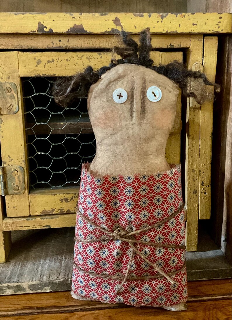May include: A handmade fabric doll with a tan face, button eyes, and dark yarn hair. The doll wears a red and blue patterned dress tied with twine. The doll is displayed in front of a distressed yellow cabinet with a wire mesh door.