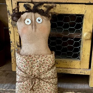 May include: A handmade doll with a tan fabric head, button eyes, and brown yarn hair. The doll's body is wrapped in a floral patterned fabric and tied with twine. The doll is a rustic, country-style craft.
