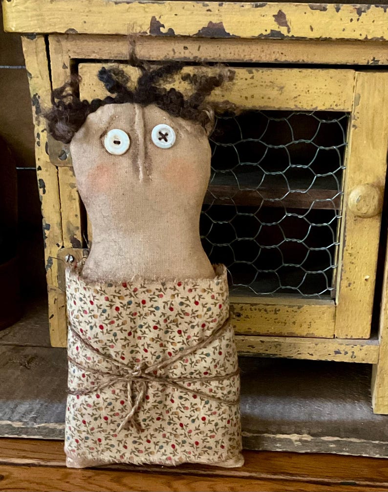May include: A handmade doll with a tan fabric head, button eyes, and brown yarn hair. The doll's body is wrapped in a floral patterned fabric and tied with twine. The doll is a rustic, country-style craft.