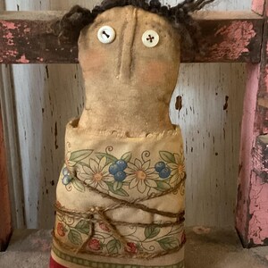 May include: A handmade doll with button eyes and dark, fuzzy hair. The doll's body is wrapped in fabric with a floral and berry pattern, tied with twine. The doll has a vintage, rustic aesthetic.