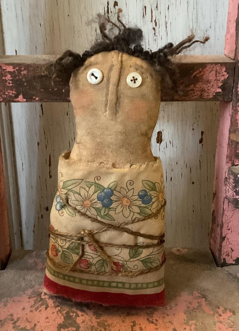 May include: A handmade doll with button eyes and dark, fuzzy hair. The doll's body is wrapped in fabric with a floral and berry pattern, tied with twine. The doll has a vintage, rustic aesthetic.