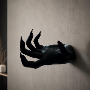 May include: A matte black wall-mounted sculpture shaped like a human hand with elongated, pointed fingers. The hand is positioned horizontally, extending outward from the wall surface, featuring a smooth finish and a distinct, claw-like posture.