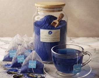 May include: A clear glass jar with a wooden lid containing vibrant blue butterfly pea flower powder and a small wooden scoop. Beside it are several pyramid-shaped tea bags labeled Butterfly Pea and a glass cup filled with blue tea.