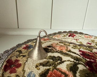 May include: A silver-toned metal candle snuffer featuring a conical bell-shaped head and a long, slender handle with a twisted design. The snuffer is positioned horizontally, showcasing its metallic finish and simple, functional construction.
