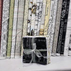 May include: A stack of folded fabric squares tied with a white ribbon, positioned in front of a row of upright fabric bolts. The fabrics feature various black and white prints, including vintage ticket stubs, cursive handwriting, botanical illustrations, and technical diagrams.
