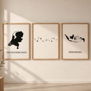 Peut inclure: Trois cadres muraux en bois clair. L'affiche de gauche présente une carte noire des Pays-Bas avec le texte en dessous. L'affiche centrale montre une ligne pointillée avec des petits cœurs. L'affiche de droite affiche une carte noire de l'Indonésie avec le texte.