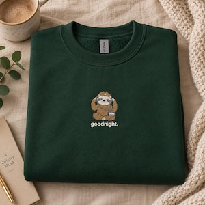 Op de afbeelding: Een donker bosgroene sweater met ronde hals voorzien van een geborduurde afbeelding van een bruine luiaard met een koptelefoon op die een kleine muziekspeler vasthoudt. Onder de luiaard staat het woord goodnight in witte kleine letters.