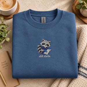May include: A blue crewneck sweatshirt featuring an embroidered graphic of a raccoon wearing blue medical scrubs and gloves, holding a sterile surgical instrument package. The text below the raccoon reads "still sterile." in small white letters.