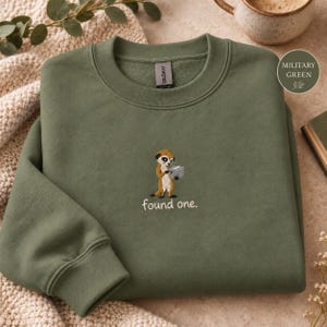 Könnte beinhalten: Ein militärgrüner Rundhals-Pullover mit einer Stickerei eines Erdmännchens, das eine kleine graue Box hält. Unter dem Erdmännchen ist der Text "found one." in Weiß gestickt. Der Pullover hat einen gerippten Kragen, Bündchen und einen Saum.
