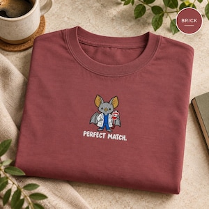 May include: A brick red crew neck t-shirt featuring an embroidered graphic of a grey bat wearing a white lab coat and blue scrubs, holding a red blood bag labelled A+. The text PERFECT MATCH. is embroidered in white below the bat graphic.