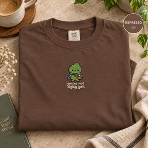 Może przedstawiać: Ciemnobrązowy t-shirt z okrągłym dekoltem i haftem przedstawiającym zielone stworzenie w fioletowej pelerynie z małą brązową torbą. Pod stworzeniem znajduje się napis "you're not dying yet". Koszulka posiada metkę marki Comfort Colors przy szyi.