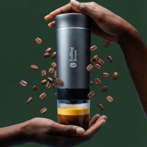 May include: A cylindrical, matte grey portable coffee maker with the text Rolling Roast printed on the side. The device features a black textured grip section and a clear bottom chamber filled with dark brown espresso, surrounded by floating coffee beans.