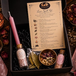 May include: A black box containing a Love Spell Kit with a pink candle, a glass bottle of rose quartz, a glass bottle of pink salt, a small bowl of dried flowers, and a plastic bag of bay leaves, alongside a printed instruction card.