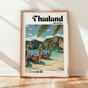 May include: A framed travel poster featuring the word Thailand in bold black text above a scenic illustration of two traditional wooden long-tail boats anchored on a sandy beach with turquoise water, palm fronds, and limestone cliffs in the background.