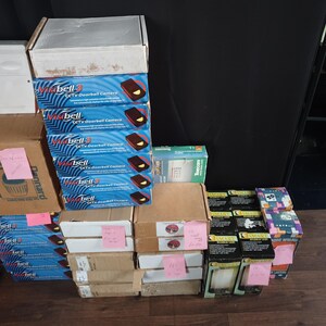 Könnte beinhalten: Eine Sammlung verschiedener verpackter Elektronikprodukte, darunter mehrere Visibell 3 CCTV-Türklingelkameras, ein weißes Sicherheitspanel und mehrere kleinere Schachteln mit der Aufschrift Smart Alarm Lamp, alle gestapelt mit kleinen rosa Klebezetteln.