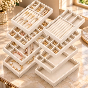 May include: A stack of beige velvet jewelry organizer trays with various compartments. The trays contain gold rings, earrings, and bracelets with floral and pearl accents. The modular design features ring rolls, square sections, and open rectangular storage areas.