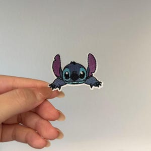 May include: An embroidered iron-on patch featuring the character Stitch from Lilo and Stitch. The patch is blue with large purple inner ears, black eyes, and a black nose, outlined in white thread against a plain background for clear visibility.