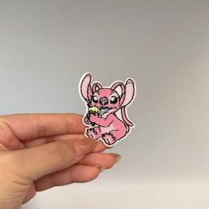 May include: An embroidered iron-on patch featuring a pink cartoon character with large ears and a nose, sitting down while holding a small yellow and blue ice cream cone. The patch has a white border and detailed stitching throughout the design.