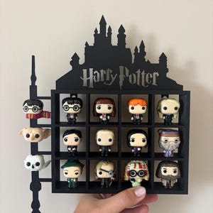Può includere: Una mensola da parete nera a forma di castello con la scritta Harry Potter. Presenta una griglia di dodici scomparti quadrati contenenti piccole statuette di personaggi, con tre teste aggiuntive fissate su un design a bacchetta laterale.