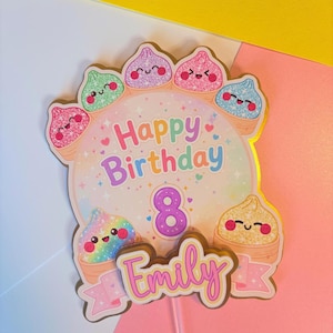 May include: A cake topper featuring a circular center with the text Happy Birthday and the number 8. Surrounding the center are several small, smiling, glittery food-themed characters. The name Emily is printed in a pink script font at the bottom.