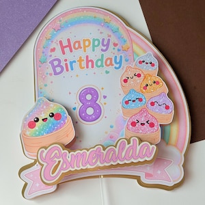 May include: A layered cake topper featuring a rainbow arch, the text Happy Birthday, the number 8, and the name Esmeralda. The design includes multiple colorful, glittery, cartoon-style dumplings with smiling faces arranged on a white stick base.