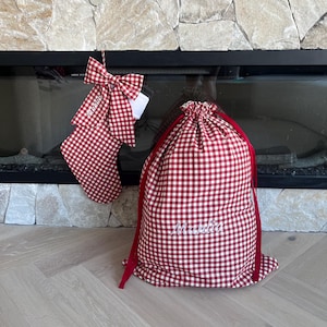 May include: A red and white gingham fabric Christmas stocking with a large matching bow and a drawstring gift sack, both featuring the embroidered name "Mathia" in white script. The items are made of a checkered textile material.